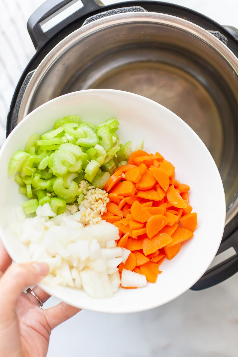 a white bowl pouring carrots, celery, onion, and garlic into an instant pot