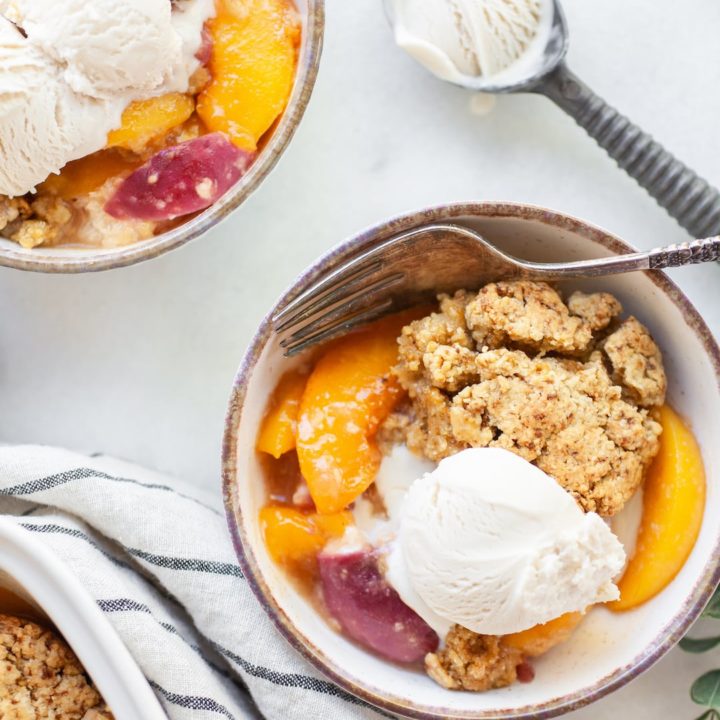 Bowl of peach cobbler and vanilla ice cream with a spoon and ice cream scoop on the side
