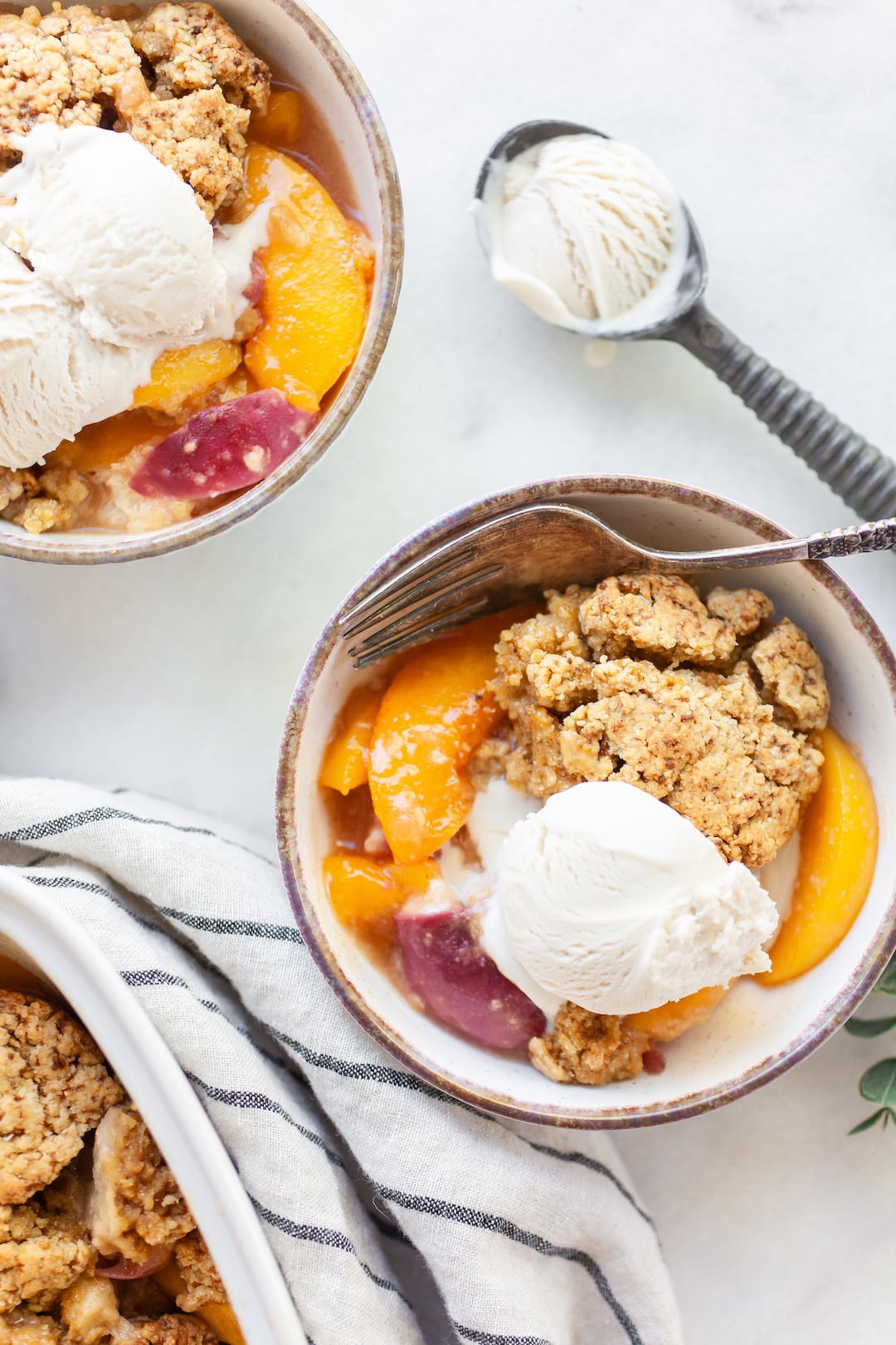 Bowl of peach cobbler and vanilla ice cream with a spoon and ice cream scoop on the side