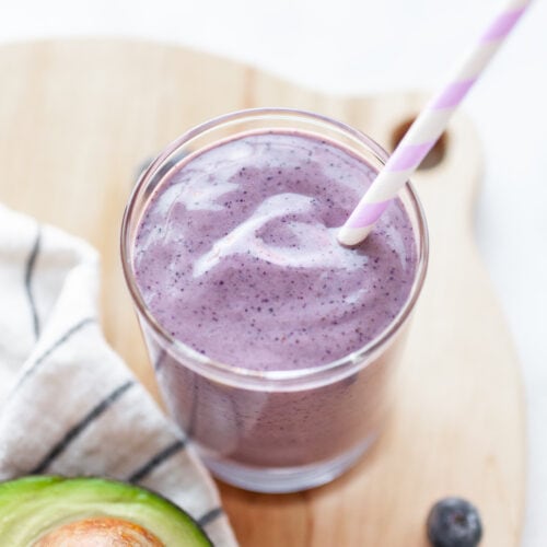 Creamy Blueberry Avocado Smoothie in a glass with a straw, avocado on the side