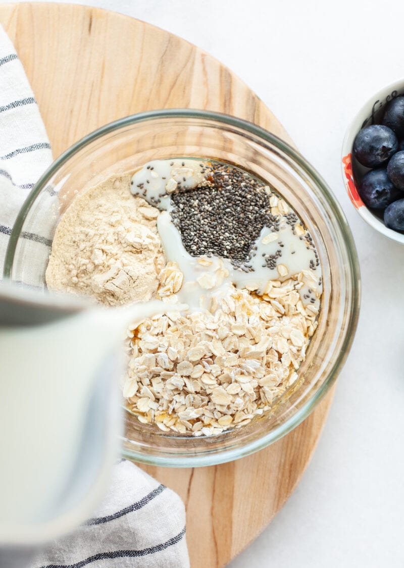 Pouring almond milk into a glass bowl with blueberry overnight oats ingredients