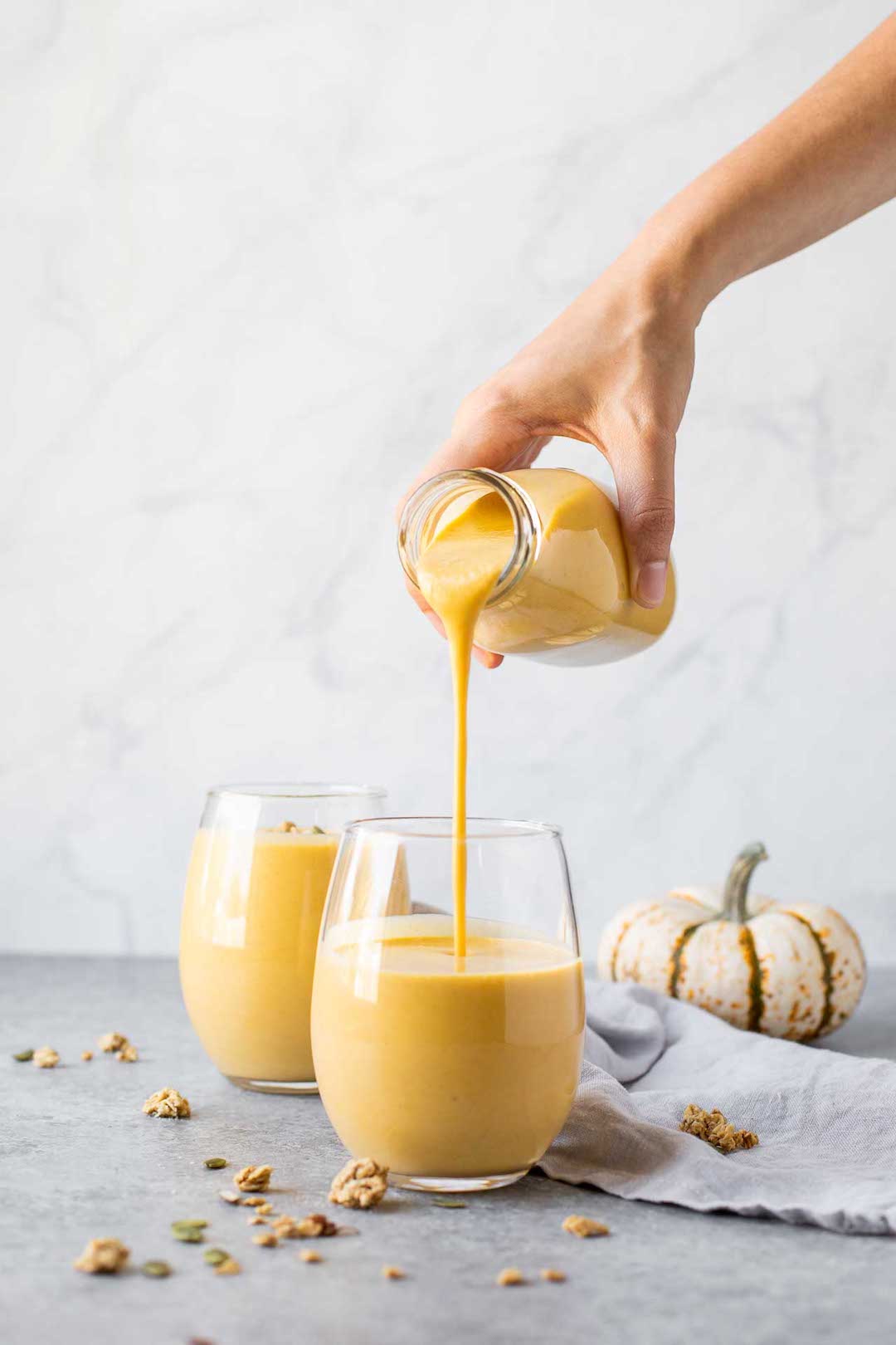 two glasses of pumpkin smoothie with a grey backdrop and smoothie being poured into one of the glasses
