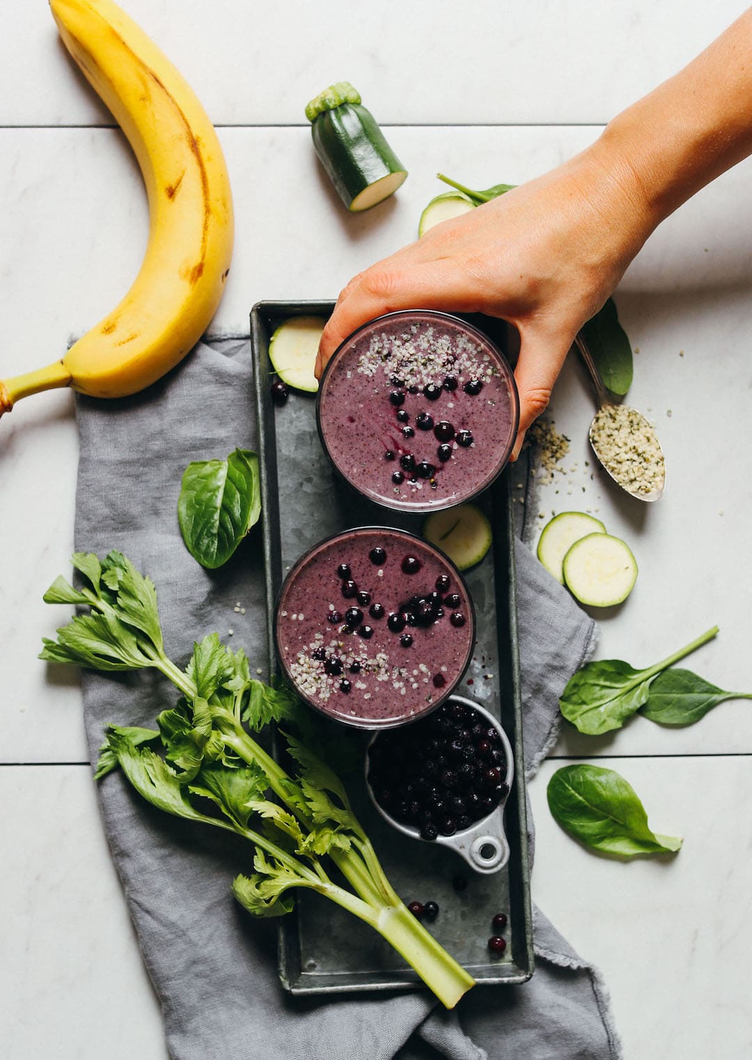 two zucchini blueberry smoothies on a tray with banana, celery, and spinach on the side