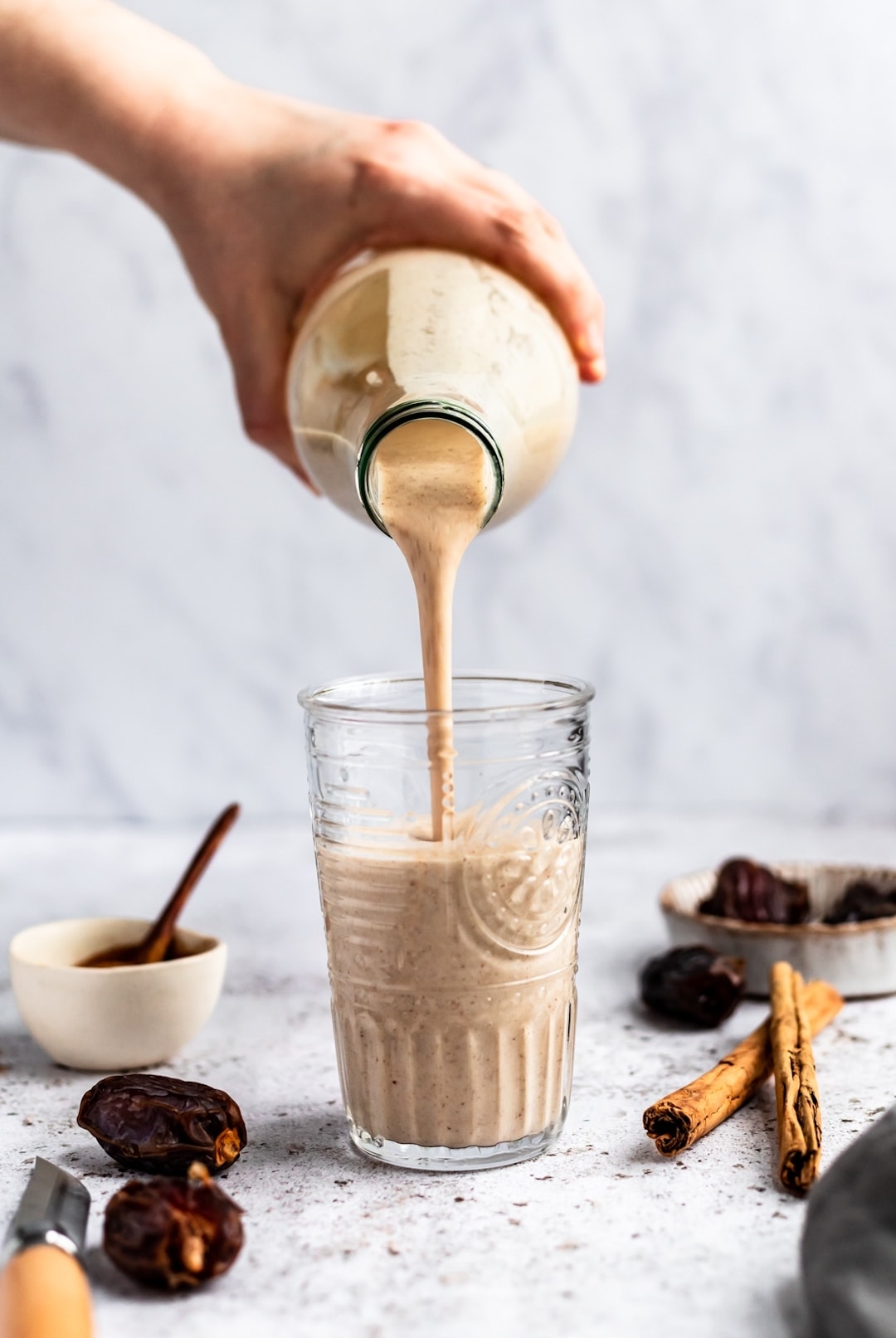Pouring snickerdoodle smoothie into a glass with dates and cinnamon sticks on the side