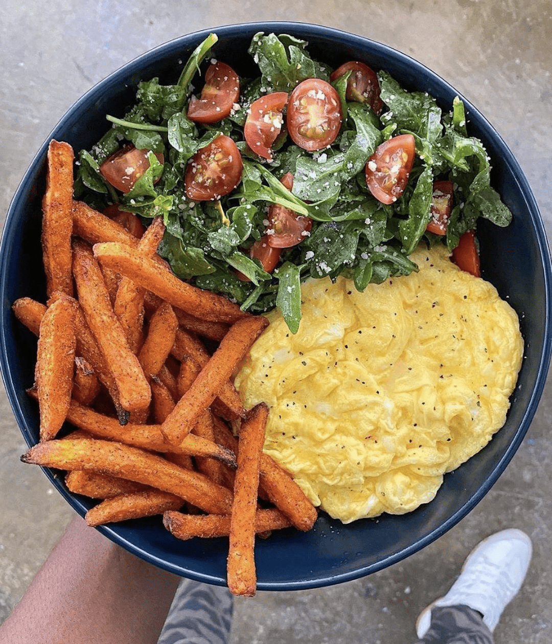 image of a hand holding a bowl with a greens salad, air fryer sweet potato fries, and scrambled eggs