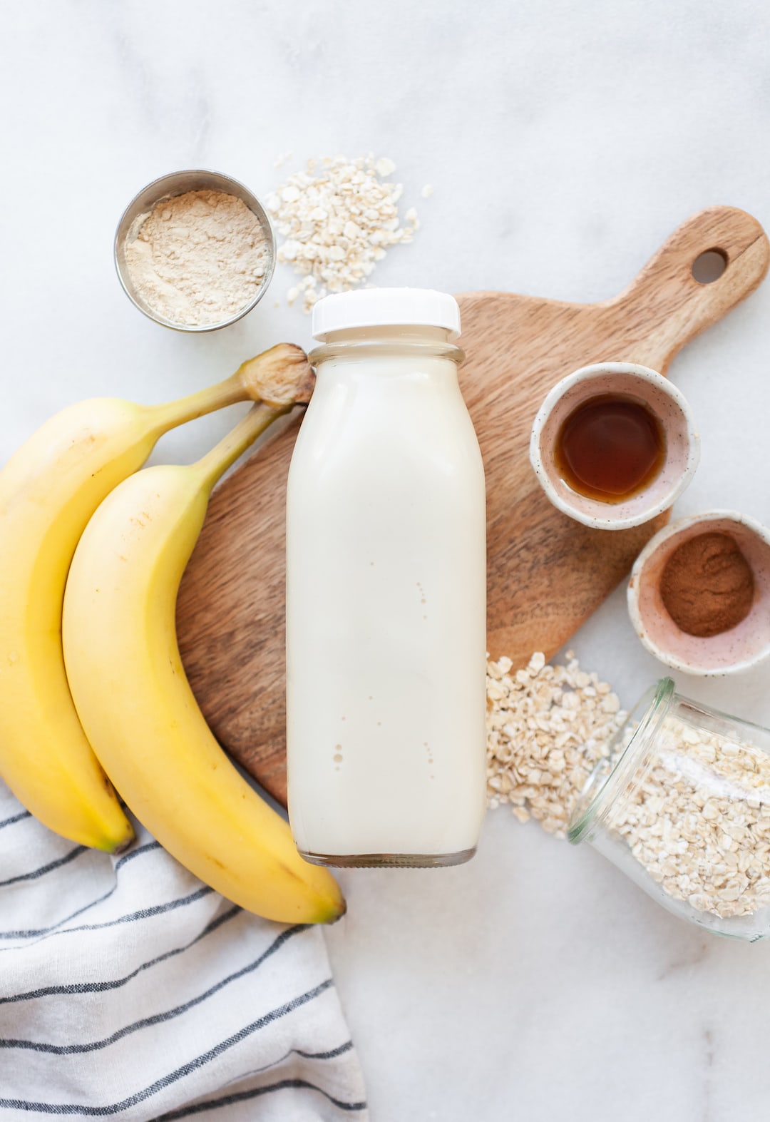 image of a glass bottle of oat milk lying sideways on a wood board with banana, oats, and cinnamon on the side