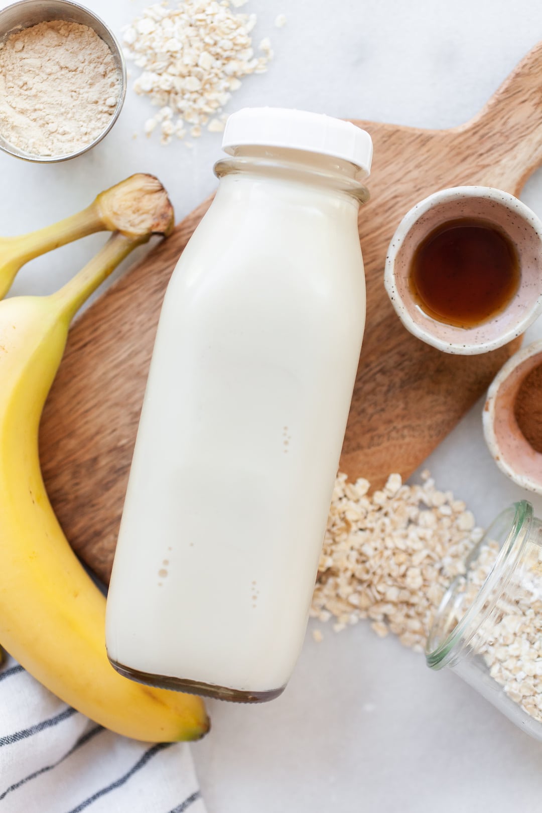 image of a glass bottle of oat milk lying sideways on a wood board with banana, oats, and cinnamon on the side