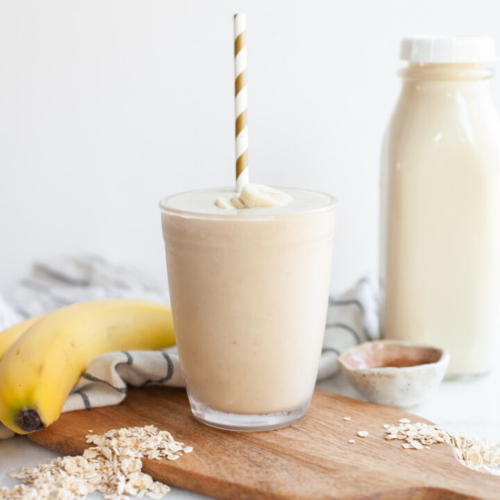 image of an oat milk smoothie on a wood board with bananas, oats, and a carafe of oat milk on the side