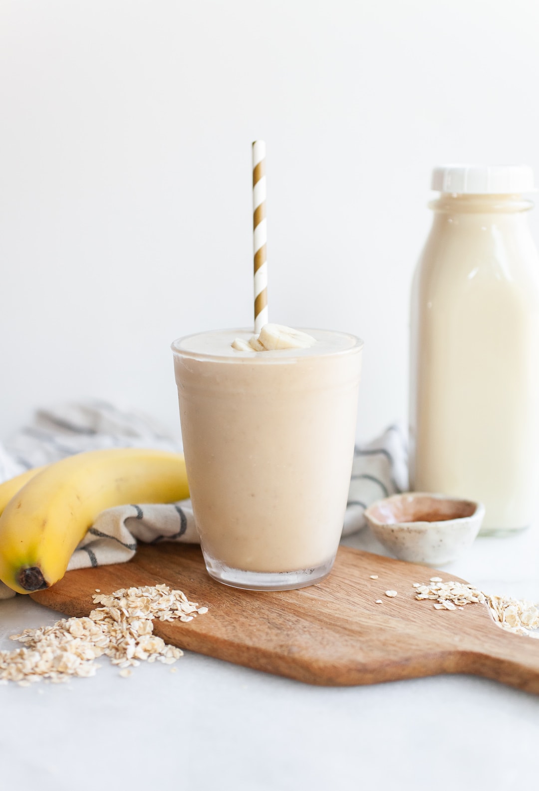 image of an oat milk smoothie on a wood board with bananas, oats, and a carafe of oat milk on the side