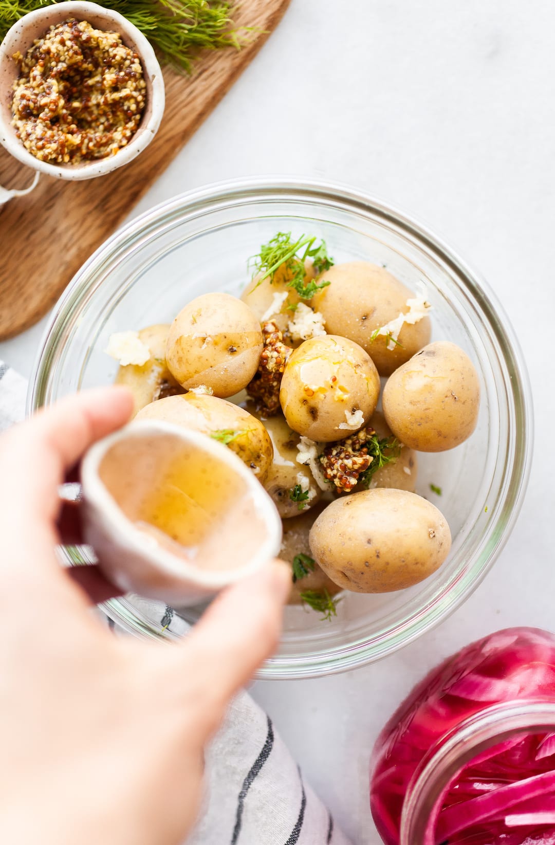 image of a bowl of boiled potatoes with herbs and dijon mustard and a hand pouring olive oil over the potatoes
