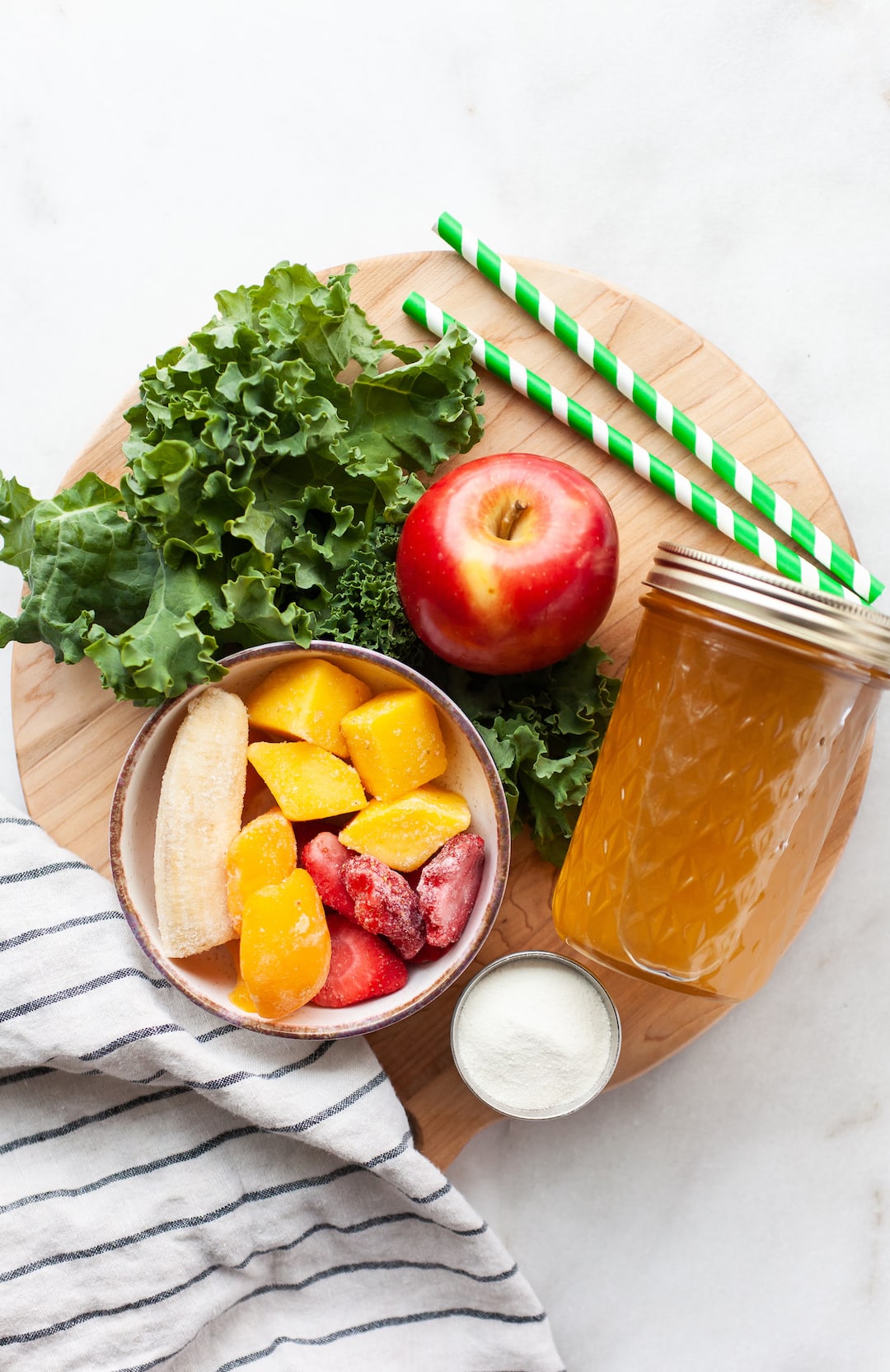 image of a wood board topped with ingredients for apple greens smoothie including frozen fruit, apple, kale, and juice