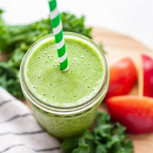image of a green smoothie surrounded by kale and red apple with a green striped straw