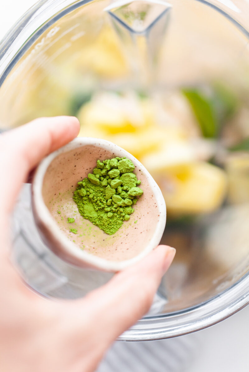 image of a small pottery bowl filled with matcha green tea powder being sprinkled into a blender canister filled with pineapple