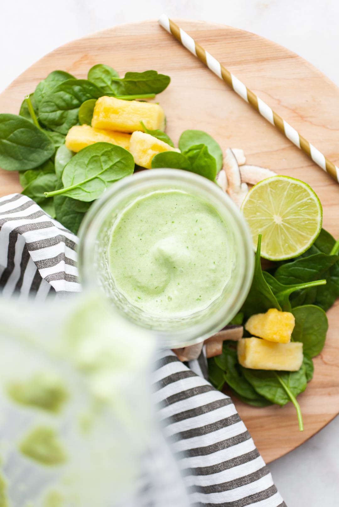 image of an overhead view of a glass jar being filled with a green smoothie poured from a blender canister and spinach, lime, and pineapple surrounding
