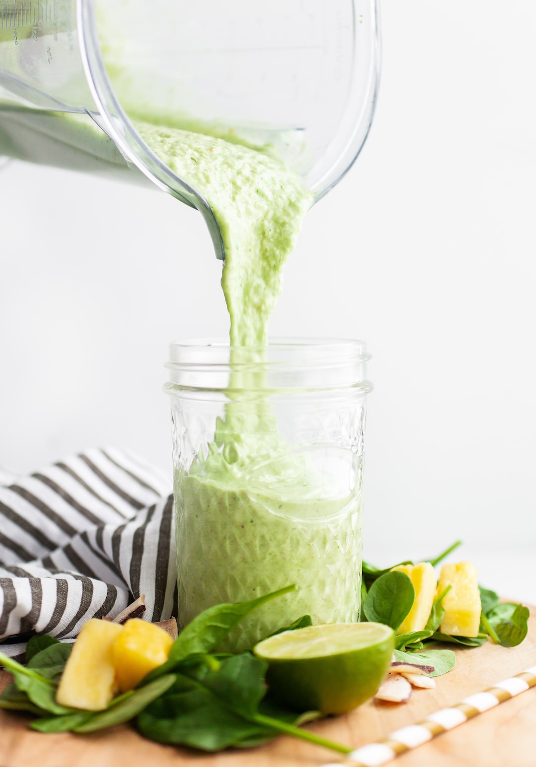 image of a green smoothie being poured from a blender into a glass jar with spinach, lime, and pineapple surrounding the jar