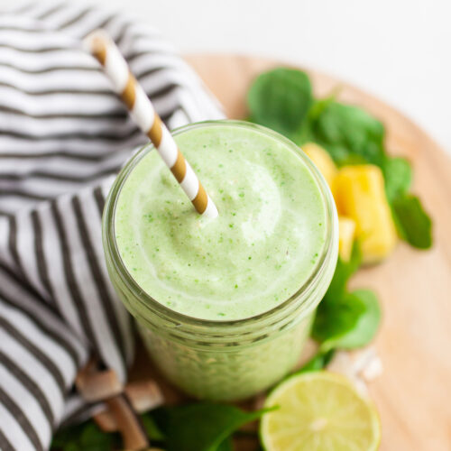 image of a glass jar filled with green smoothie on a wood board with pineapple and spinach on the side and a striped golden straw in the smoothie