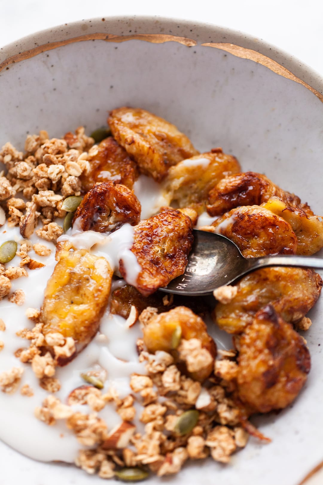 close up image of a pottery bowl filled with air fryer banana, yogurt, and granola and a spoon scooping out of the middle